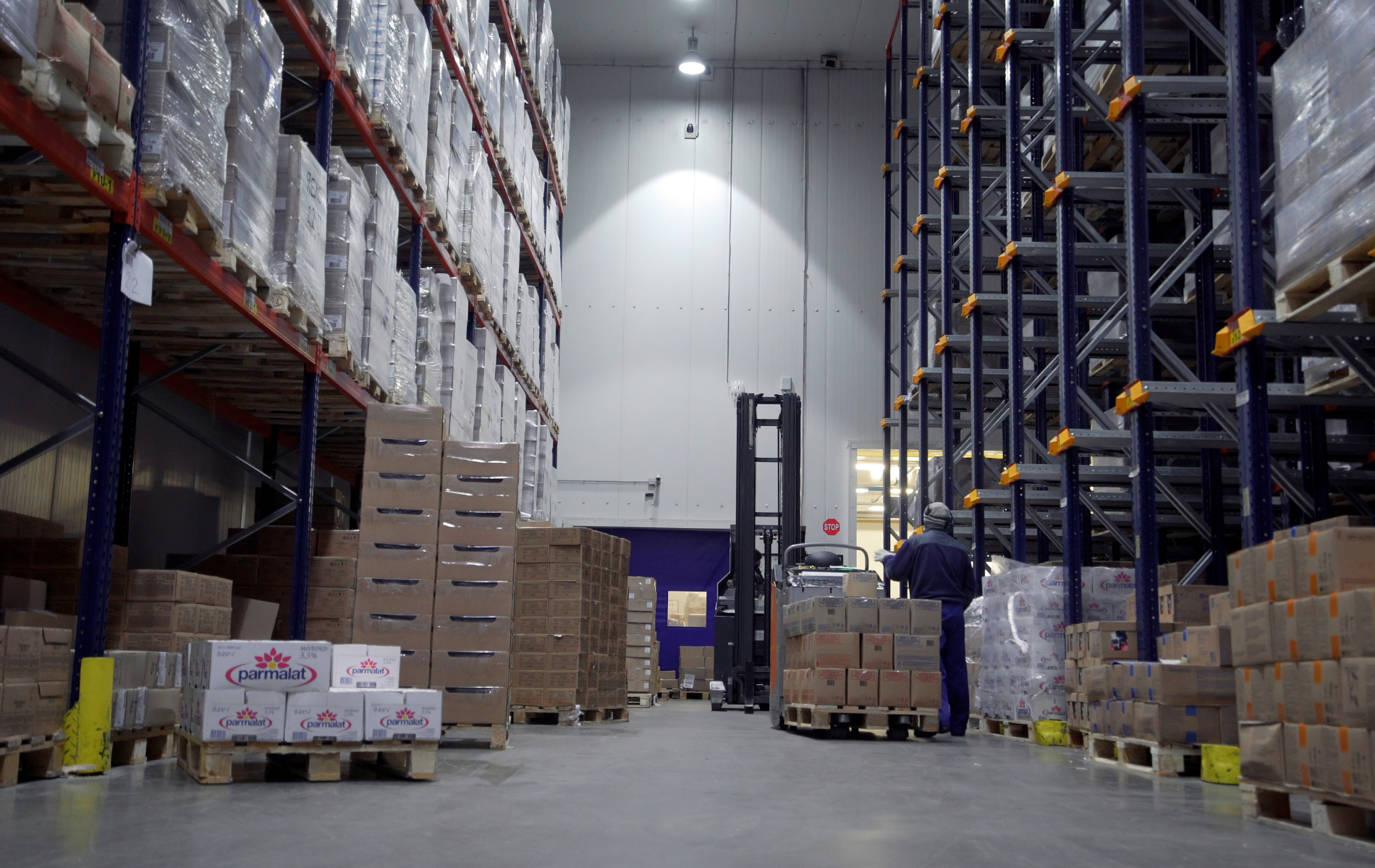 A person operates a forklift in a spacious, dimly lit warehouse filled with tall shelving units stacked with goods.