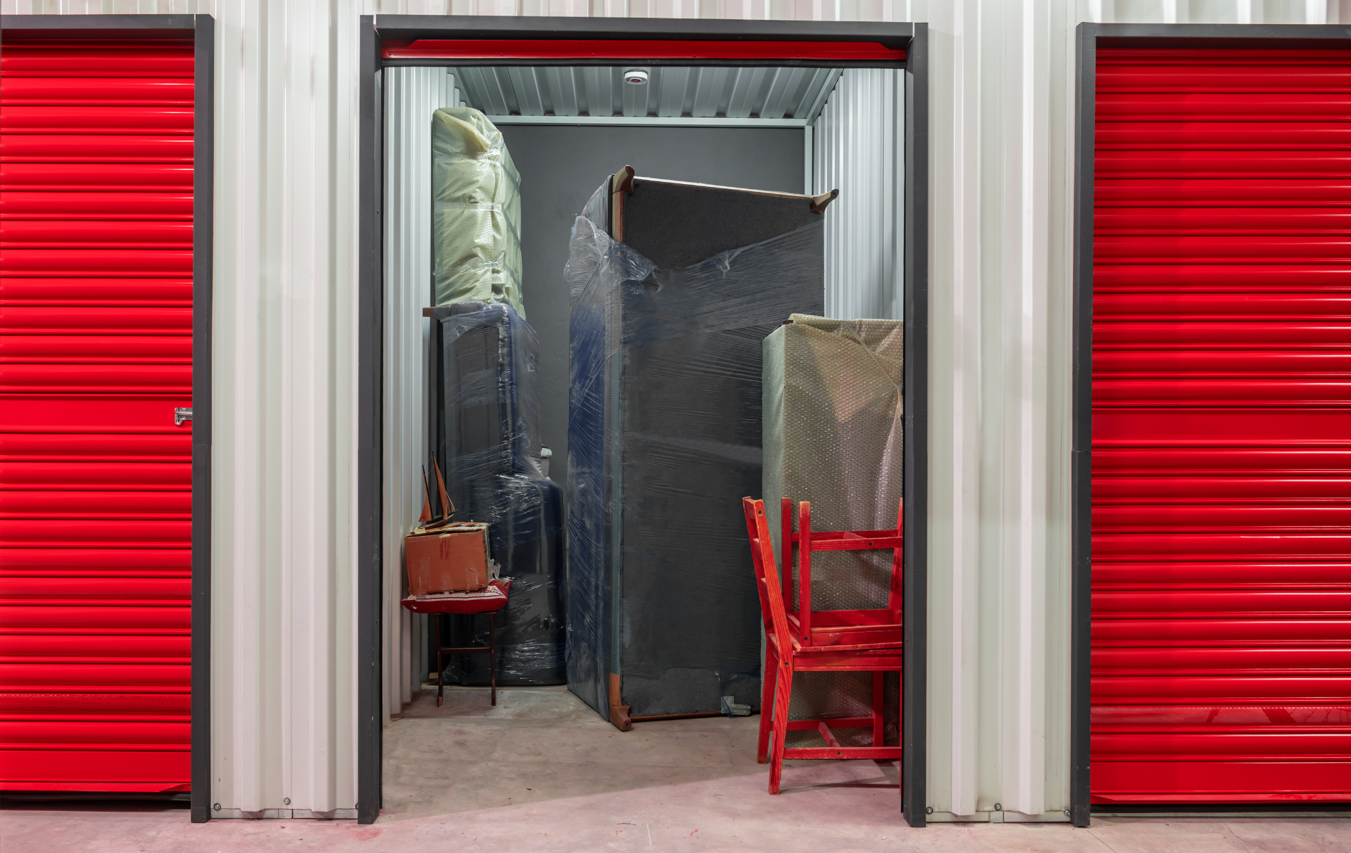 An open storage unit with red corrugated doors on both sides, containing furniture wrapped in plastic and a red chair.