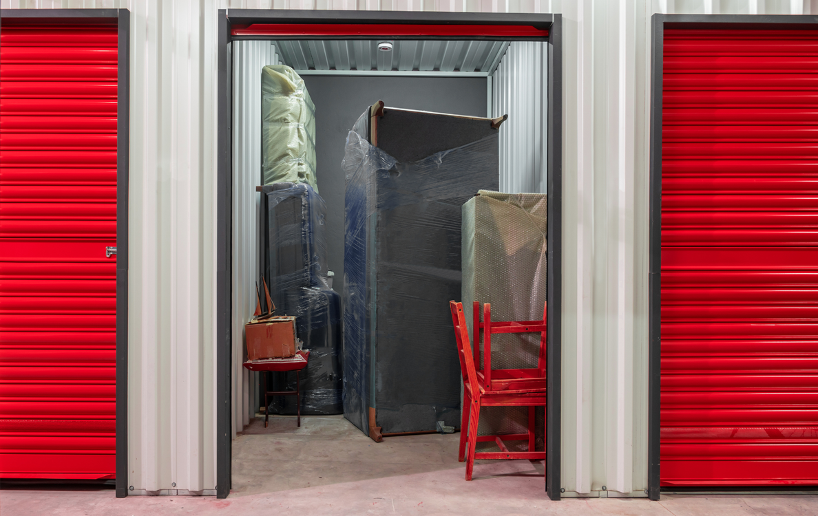 An open storage unit with red corrugated doors on both sides, containing furniture wrapped in plastic and a red chair.