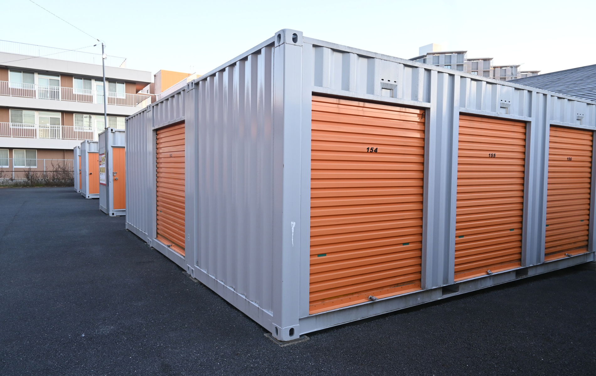 A row of grey storage containers with orange roll-up doors, set on an asphalt lot near a building.
