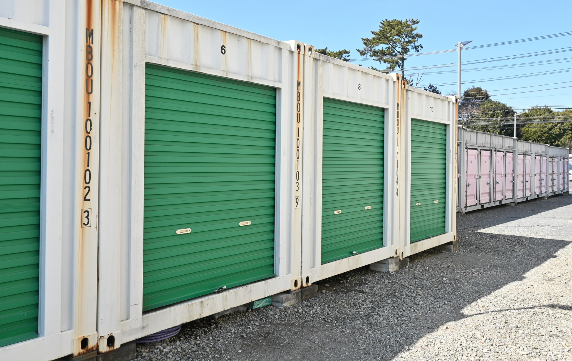 A row of white shipping container storage units with green roll-up doors, set on a gravel lot under a clear blue sky.
