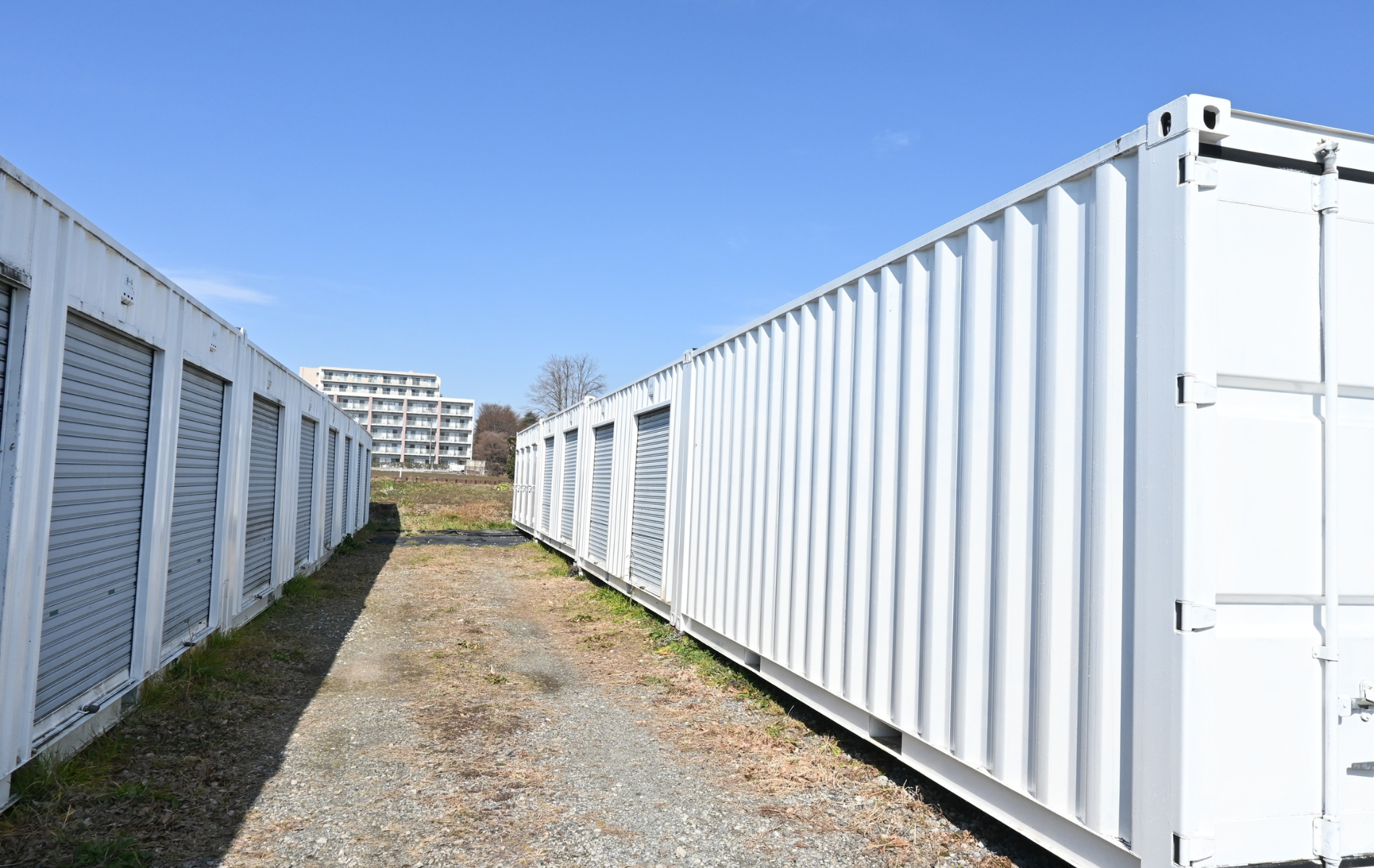 Two rows of white storage containers face each other on a gravel path under a clear blue sky.