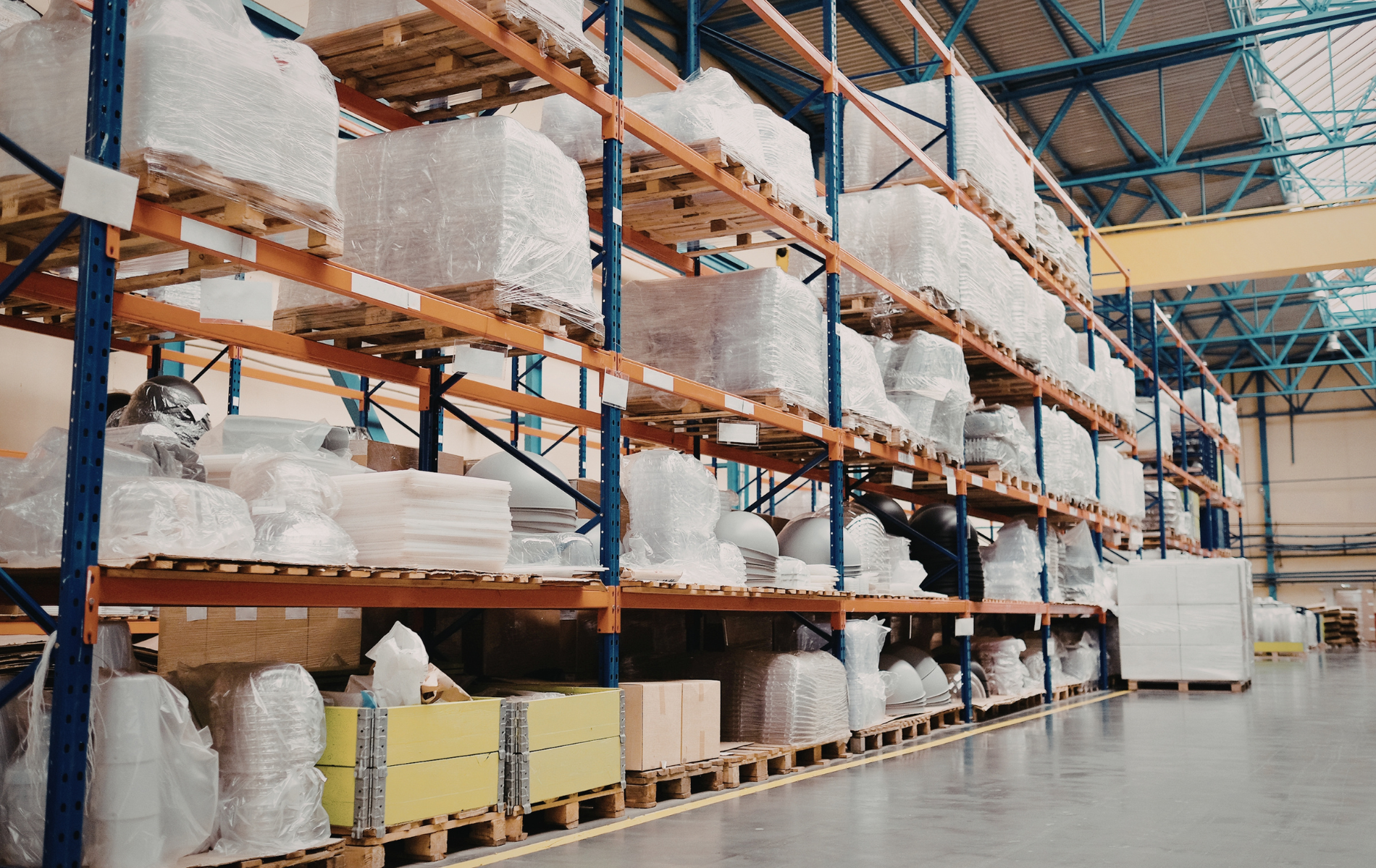 Tall industrial metal shelving units in a warehouse, stacked with palletized goods wrapped in white plastic.