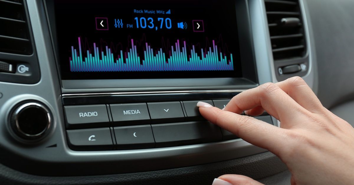 A manicured hand and holding down a button on an infotainment system in a car; the screen has “FM 103,70” lit up on it.