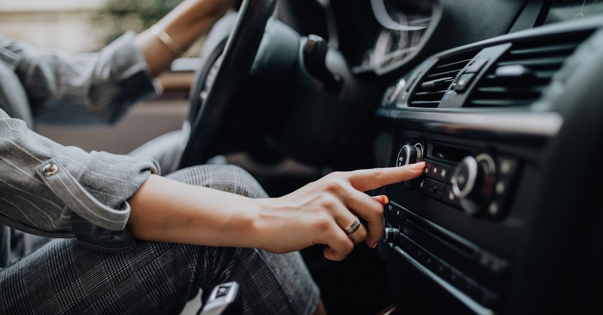 A person has a hand on the steering wheel and one on a button on a car’s dashboard; the interior of the car is all black.
