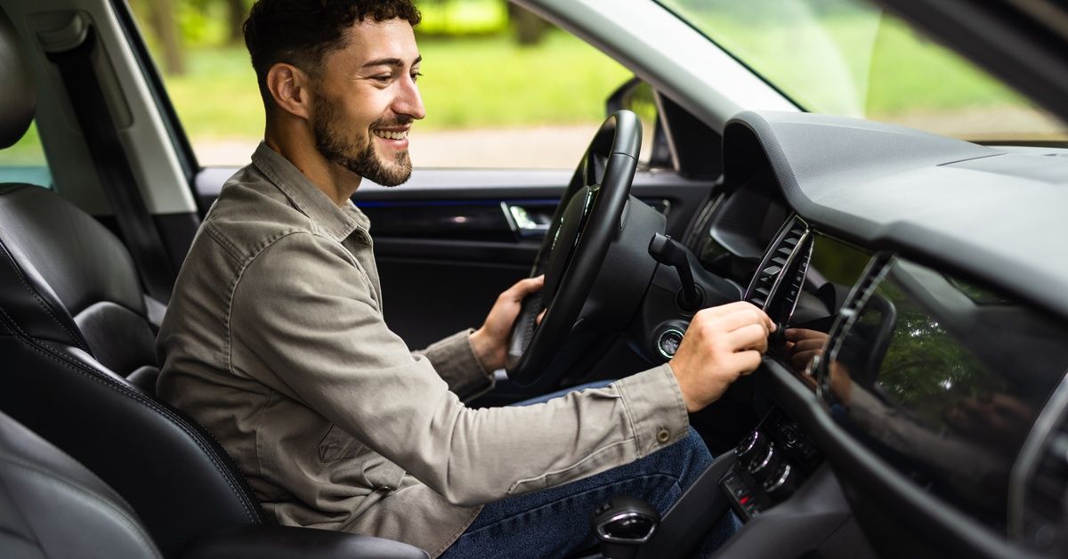 A young man smiling and wearing a long sleeve shirt smiles and sits inside of a car with a hand on t