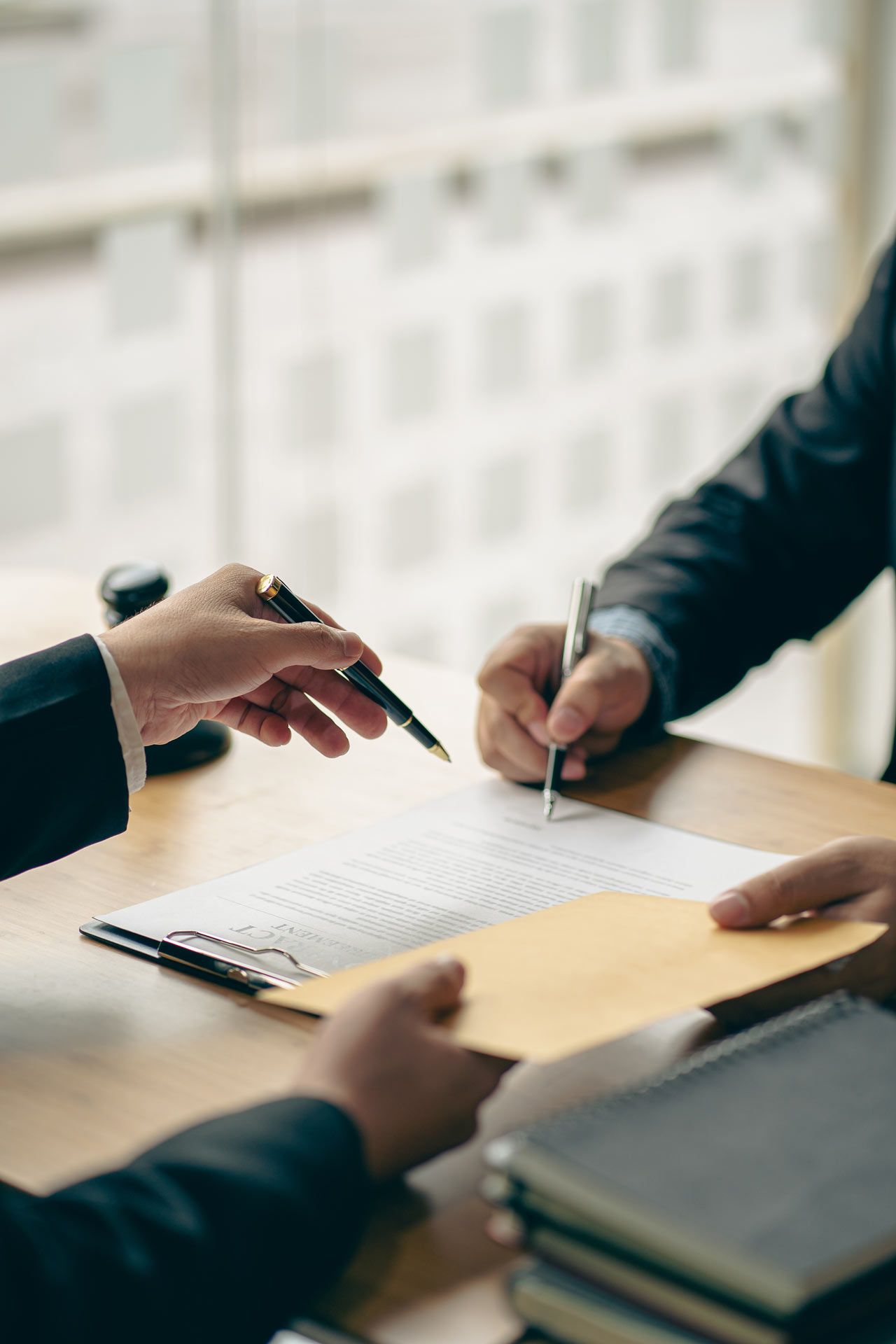 Two people in suits signing documents at a desk, one holding out a pen, the other holding an envelope.