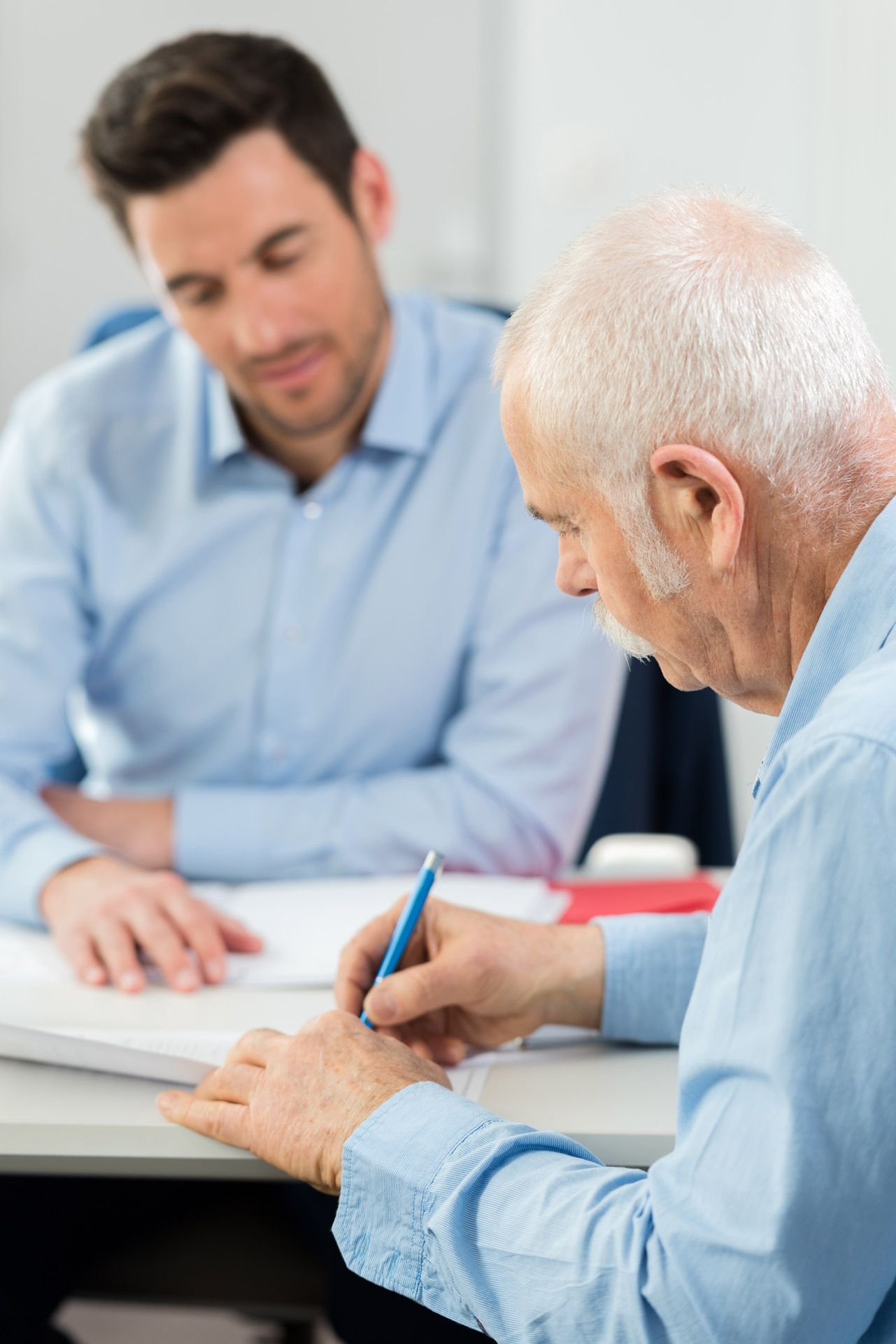Senior signing a document, guided by a younger man in an office setting.