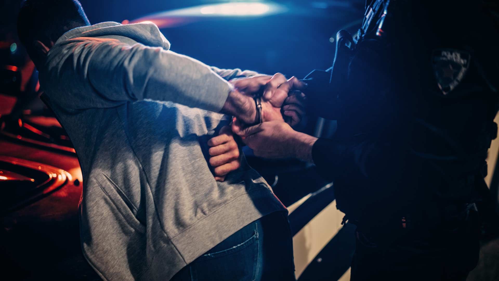 Police officer handcuffs a person against a car, bright blue and red flashing lights.