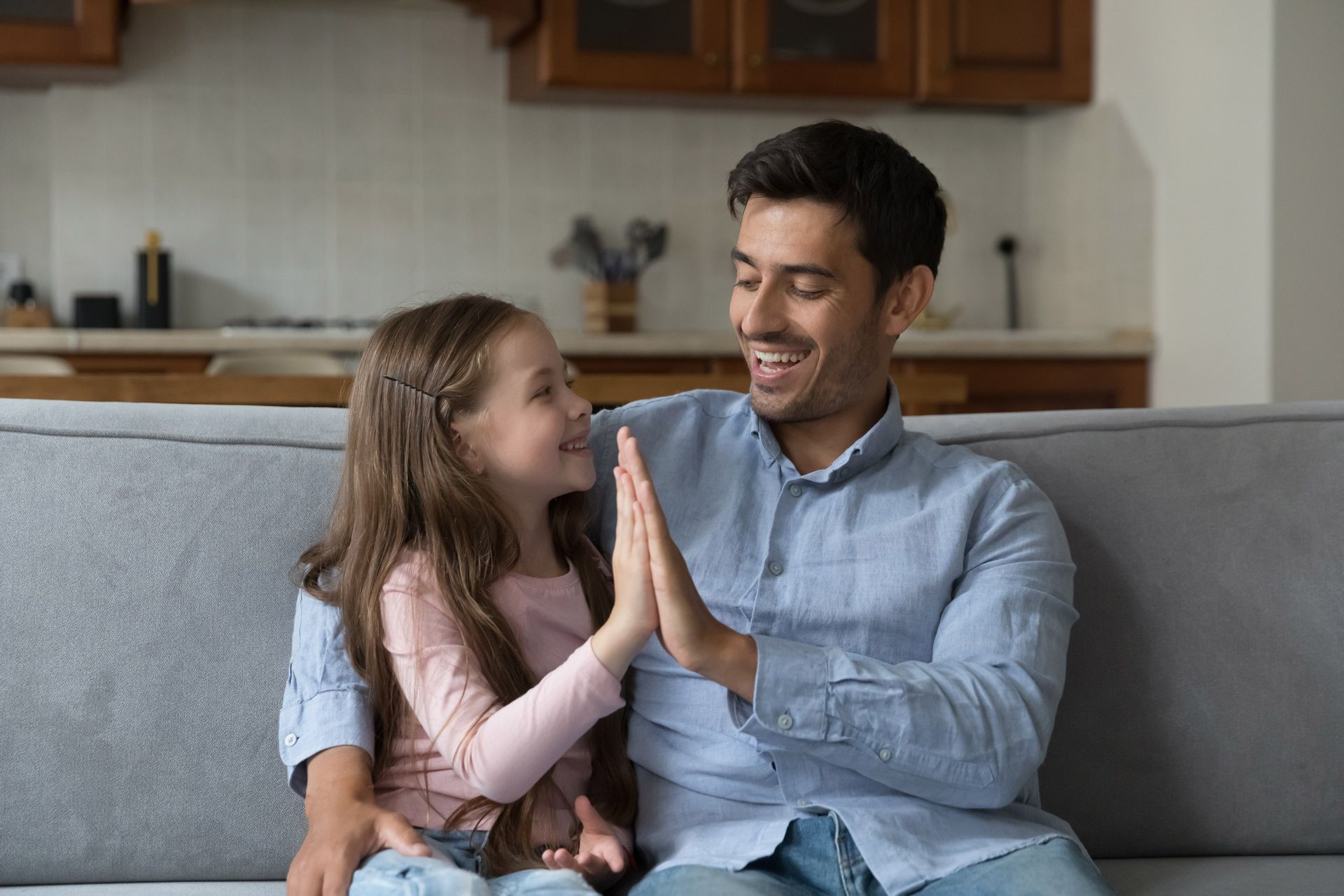 Father and daughter high-fiving on a couch, smiling. They're in a living room with a kitchen in the background.
