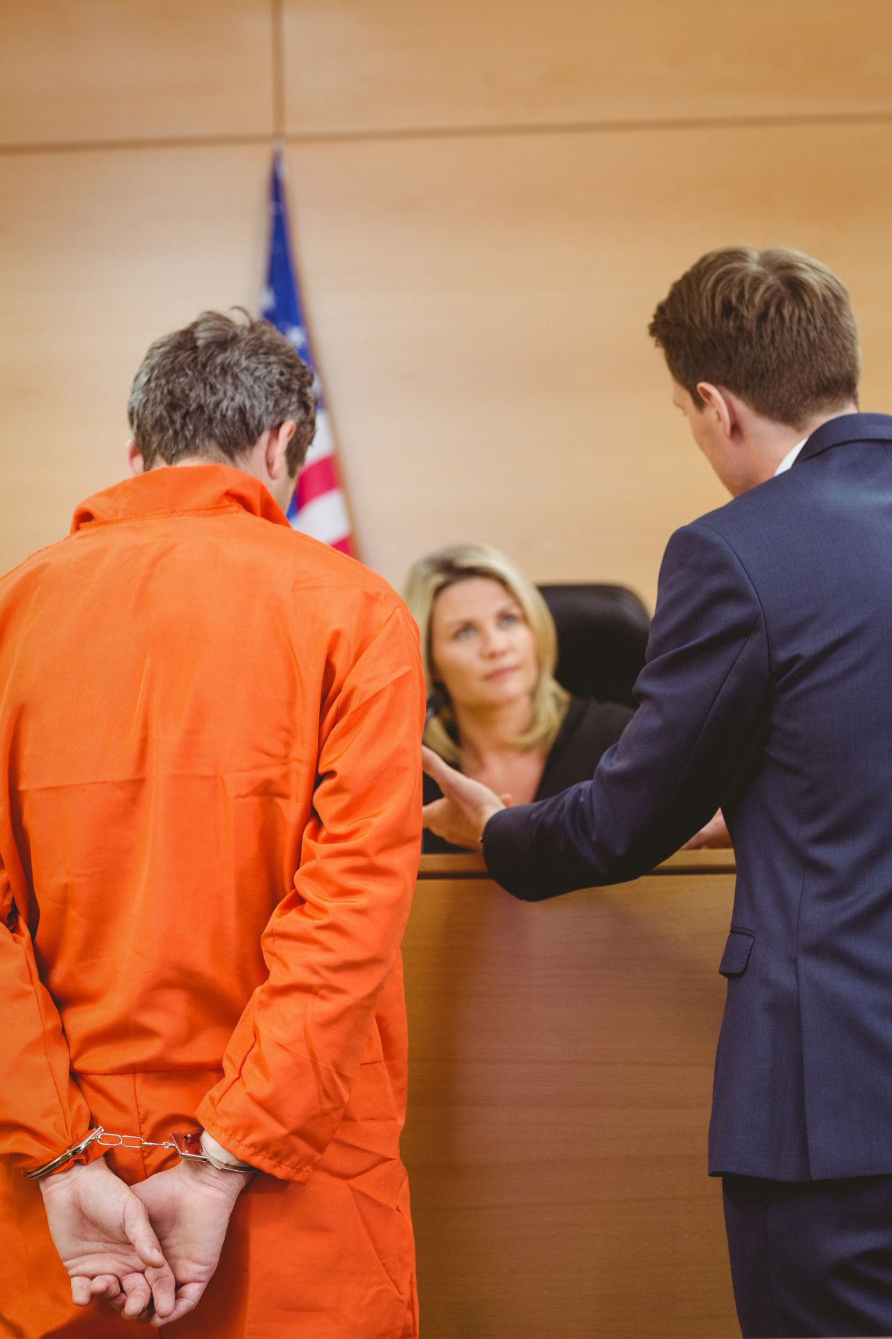 A man in handcuffs and orange jumpsuit stands before a judge and lawyer in a courtroom.