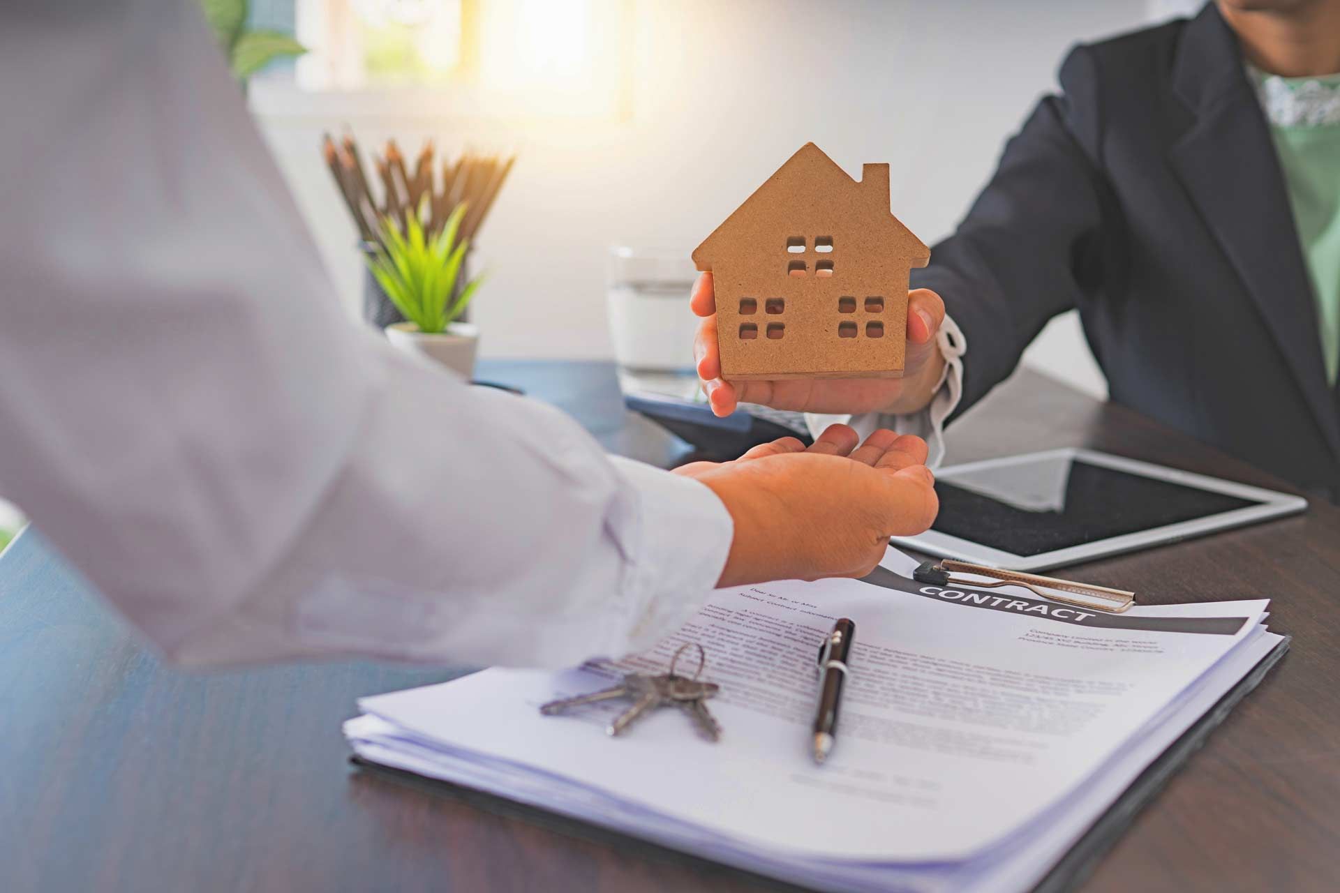 Person handing a model house to another person over paperwork and keys on a desk.