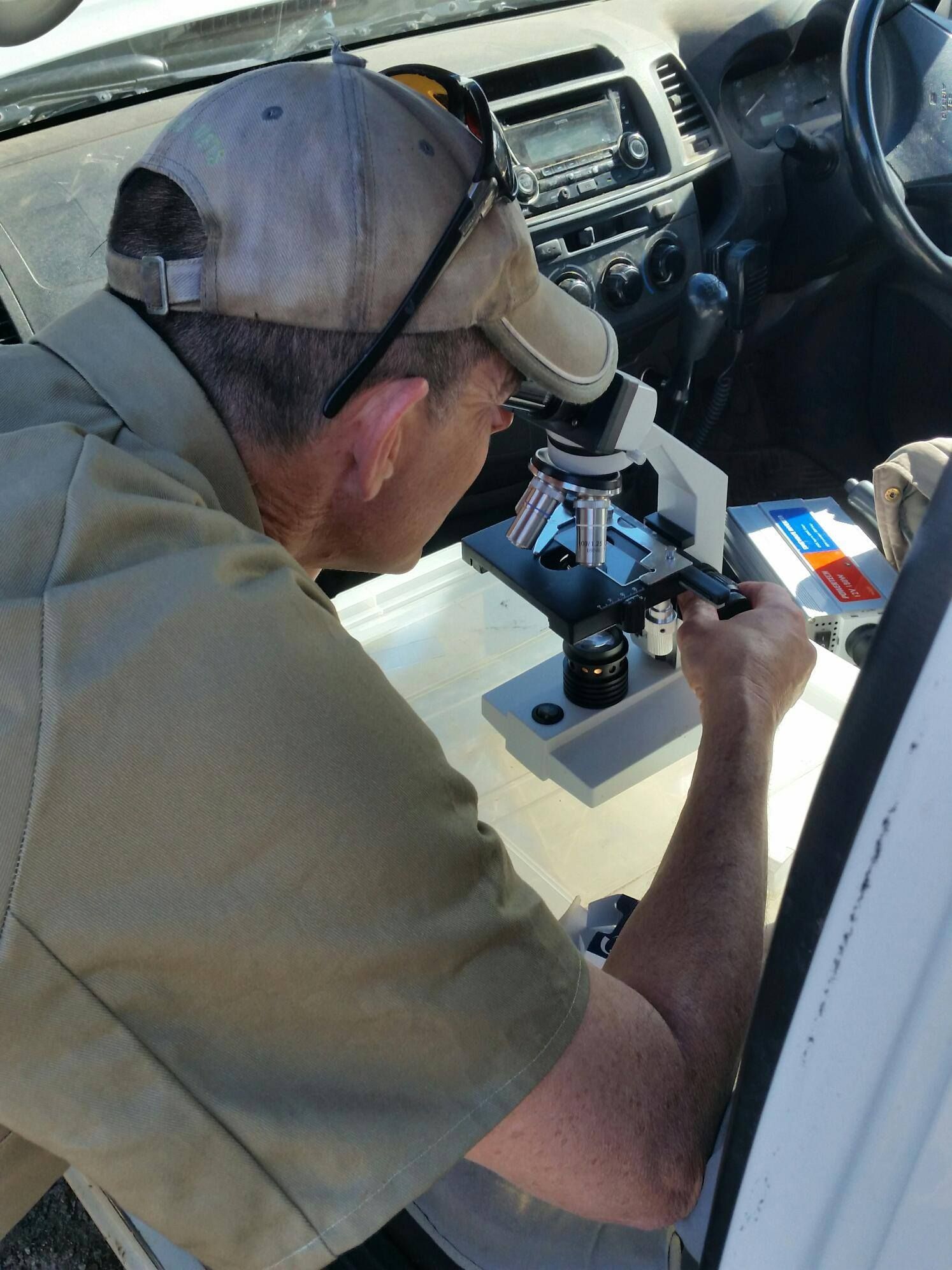 Man looking into a microscope inside a vehicle. He wears a cap and tan shirt.