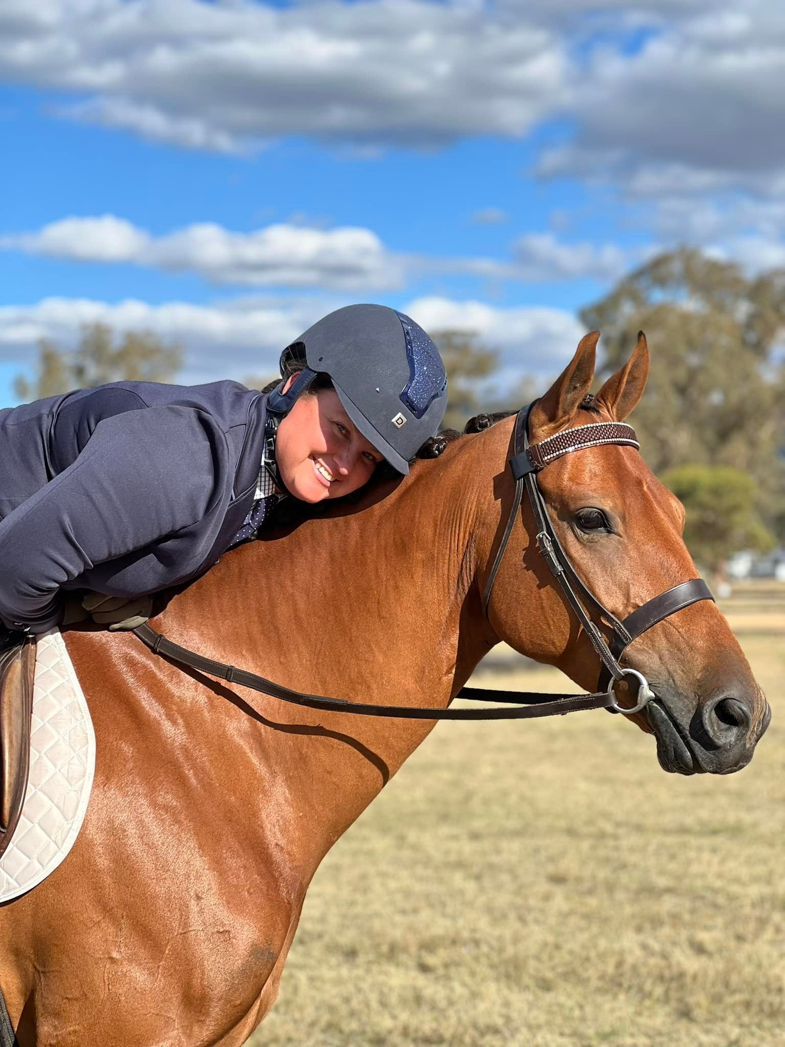 Woman in riding gear smiling, hugging a brown horse in a field under a blue sky.