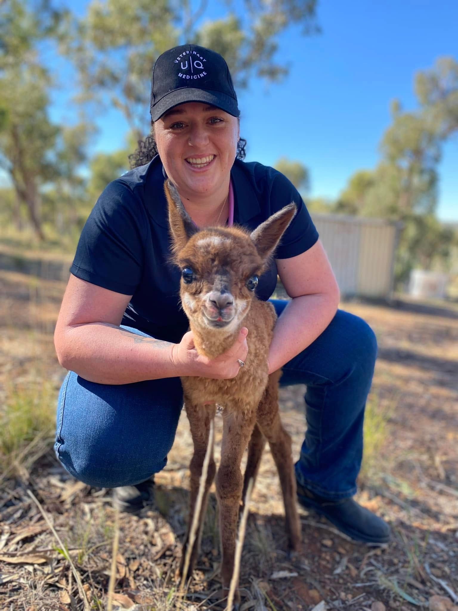 Woman kneeling, holding a young brown kangaroo in an outdoor setting, smiling at the camera.