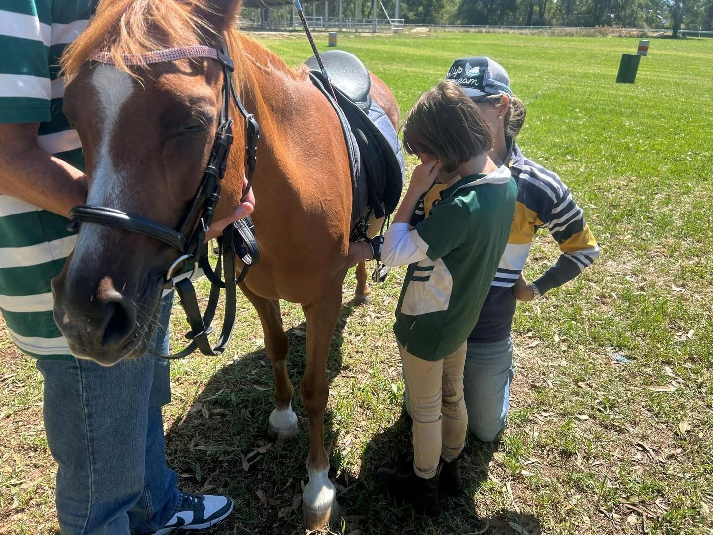 A brown horse is saddled with two children and an adult standing beside it outdoors.