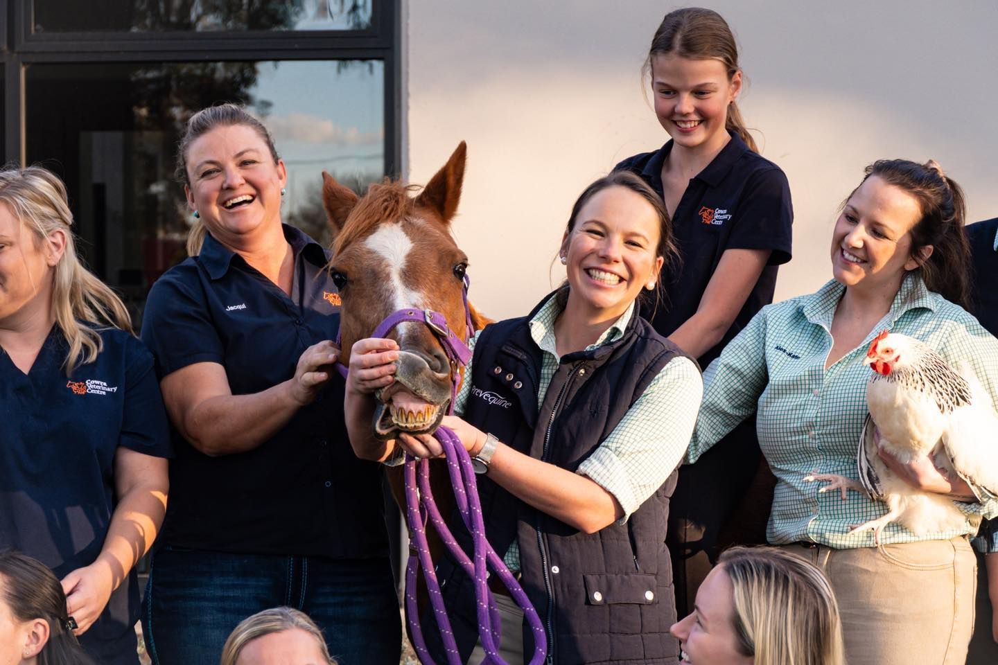 Group of women smiling with a foal and a chicken outside.