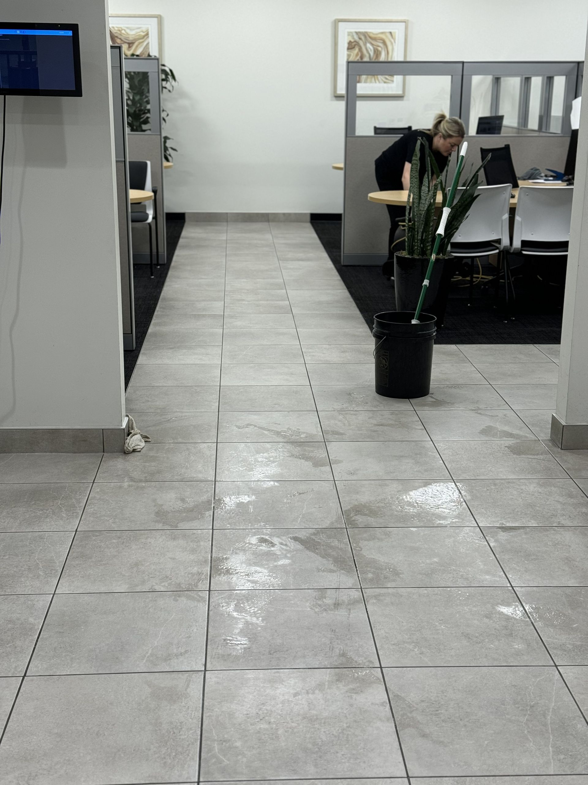 An office hallway with gray tiled floor. A person near a potted plant and office cubicles.