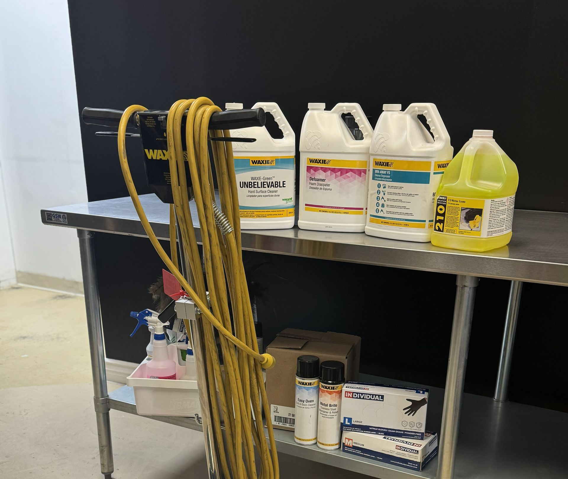 Metal table with cleaning supplies: spray bottles, chemicals, and a power cord against a black backdrop.