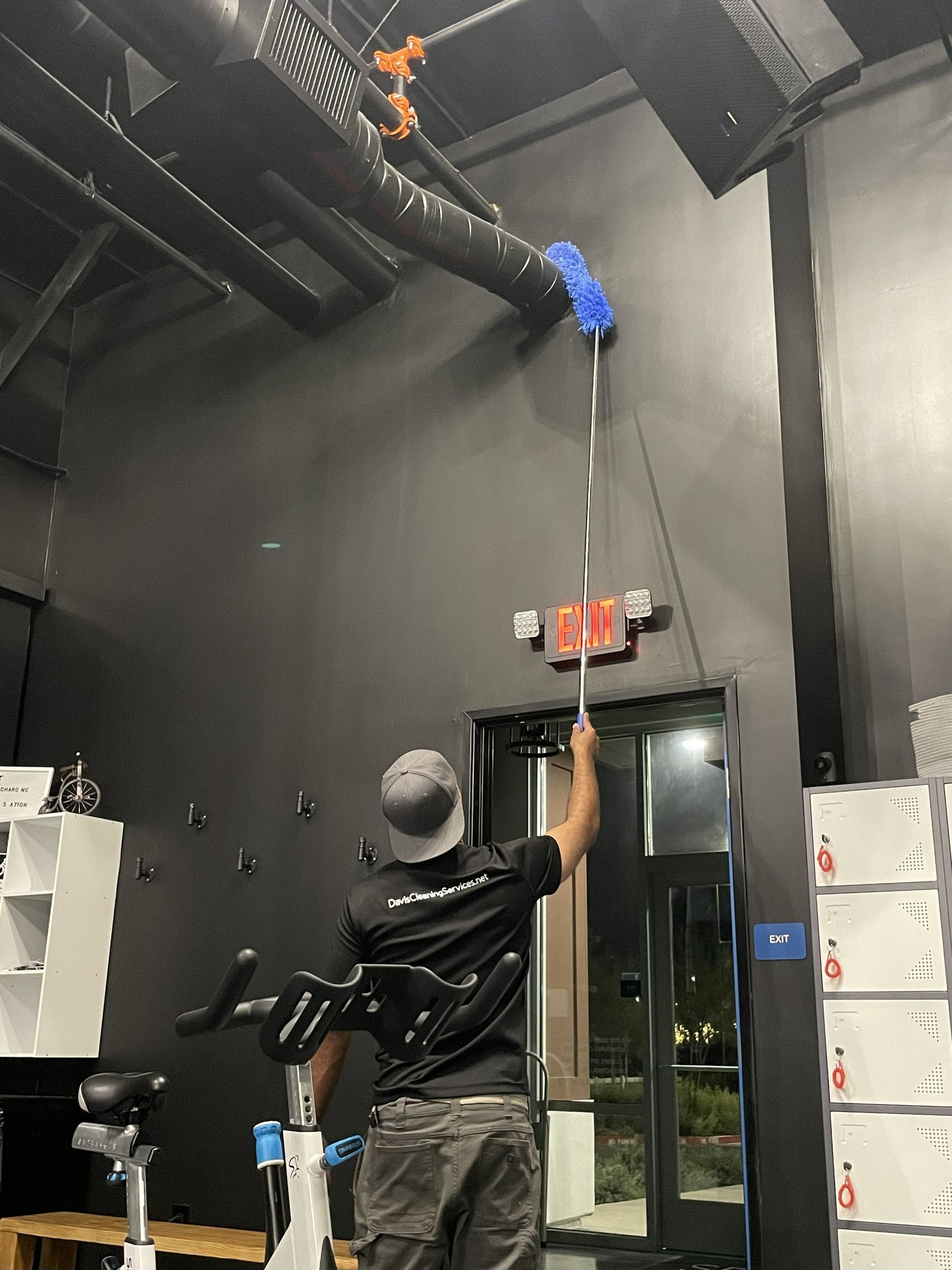 Person dusts high wall and pipes with a long-handled duster near an exit door in a dark room.