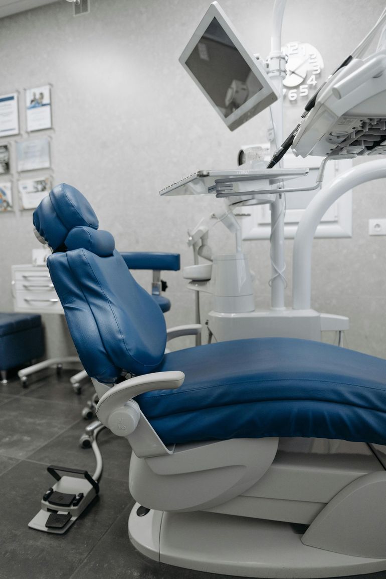 Blue dental chair in a dentist's office with attached equipment; a foot pedal rests on the floor.