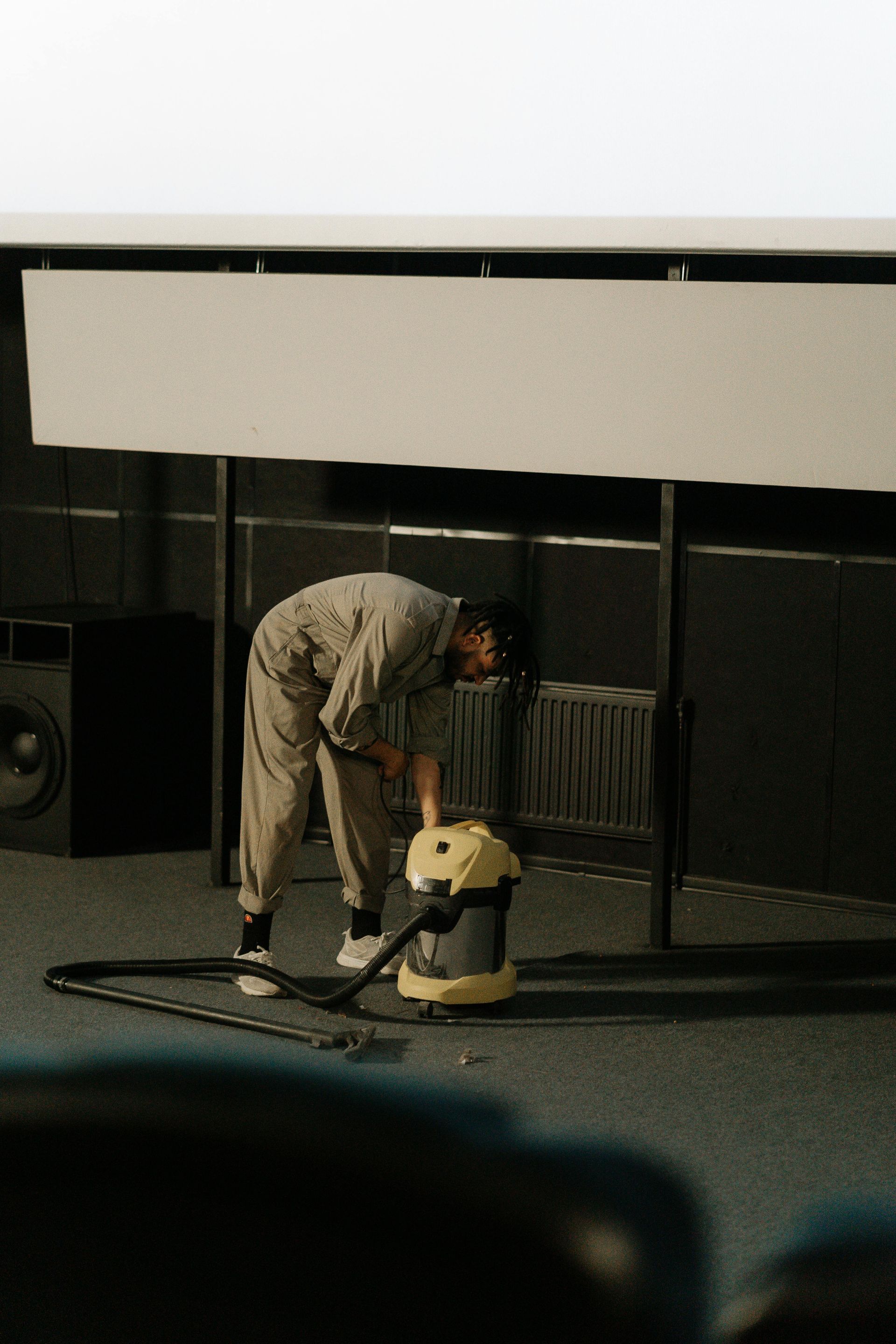 Person in coveralls vacuums a room with a large screen, near a speaker and metal bars.