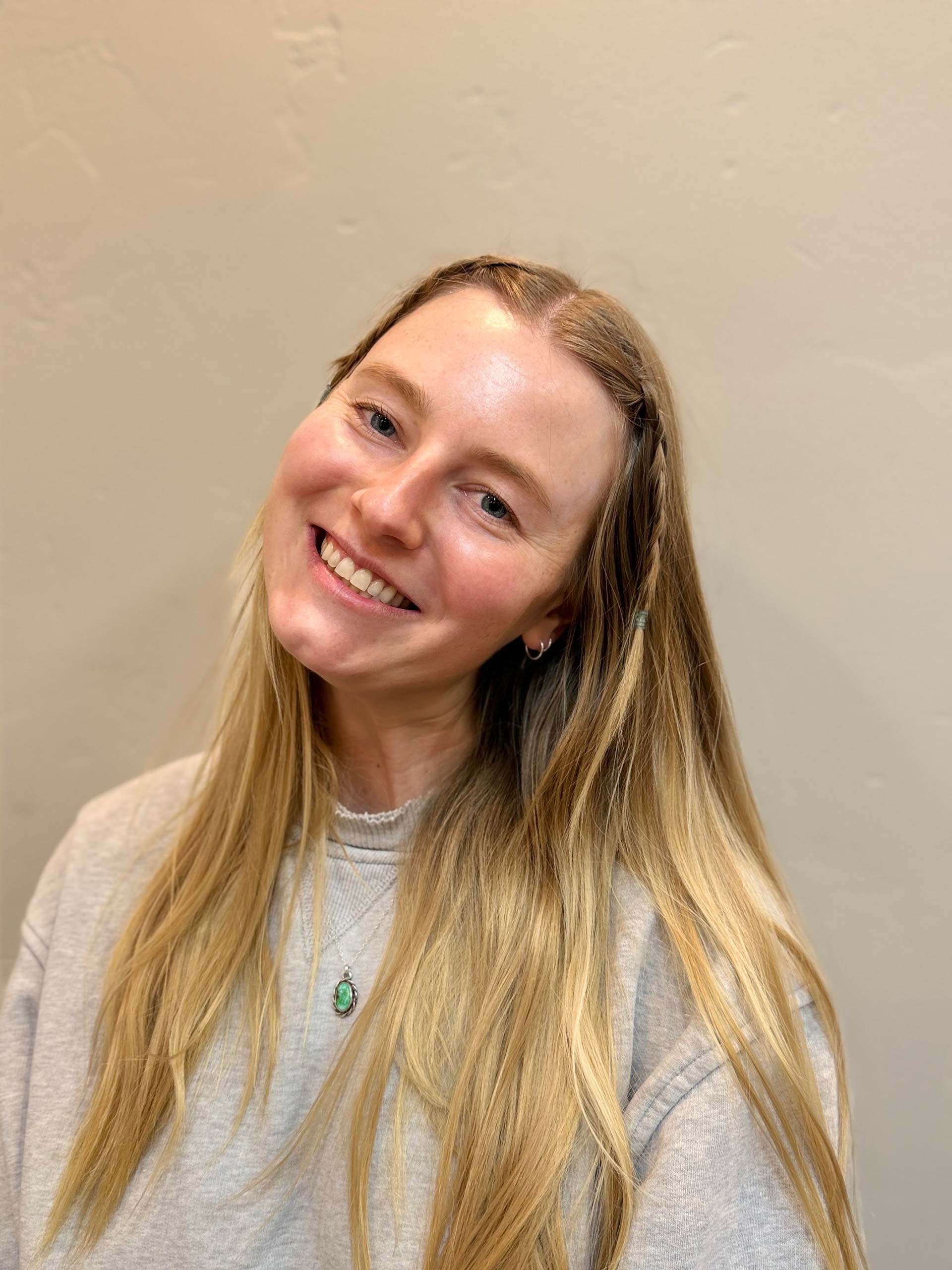 Woman with long blonde hair smiles, wearing a gray sweatshirt, and a teal pendant.