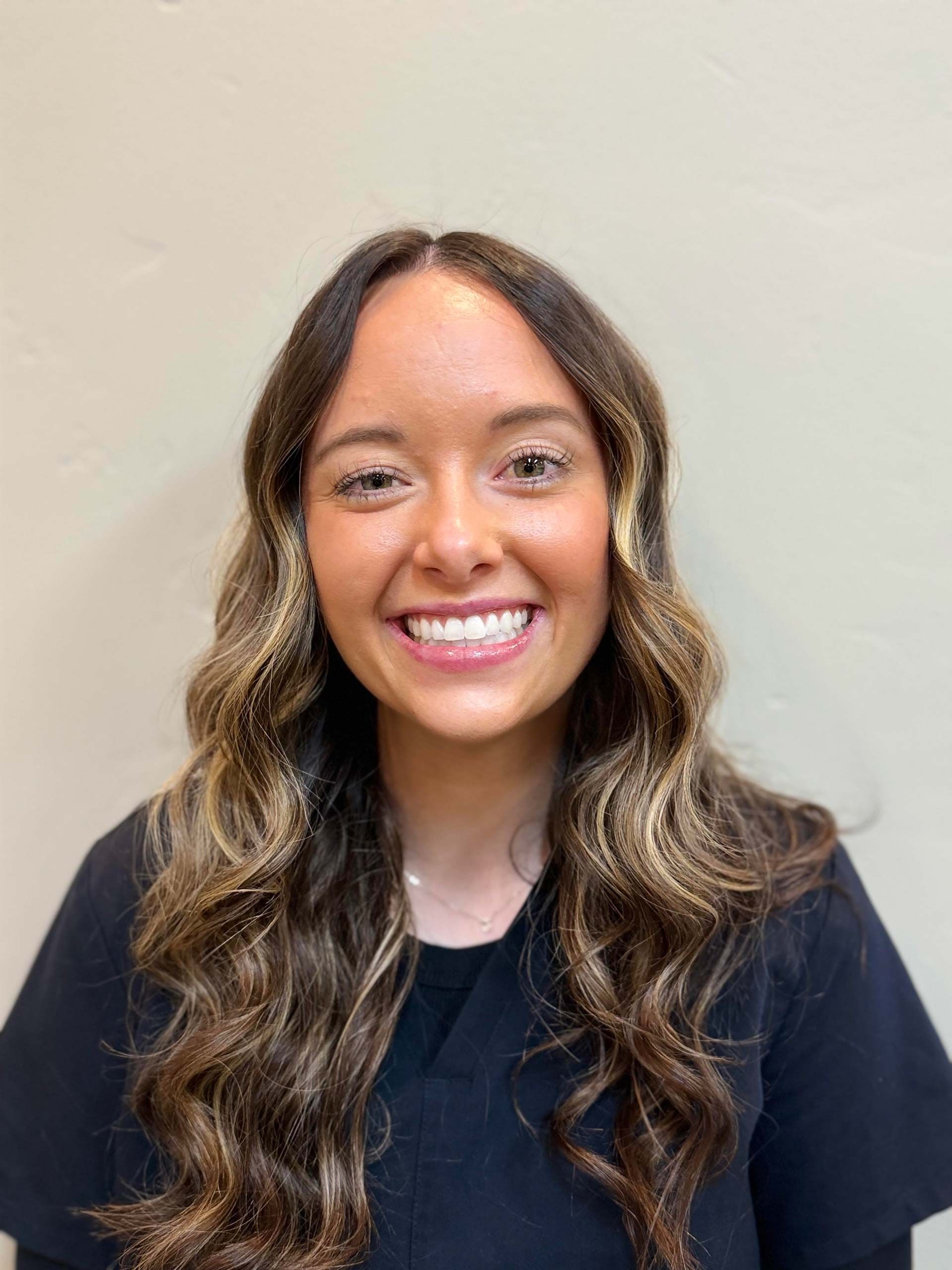 Woman with long, wavy hair smiles broadly, wearing a black top against a light-colored background.
