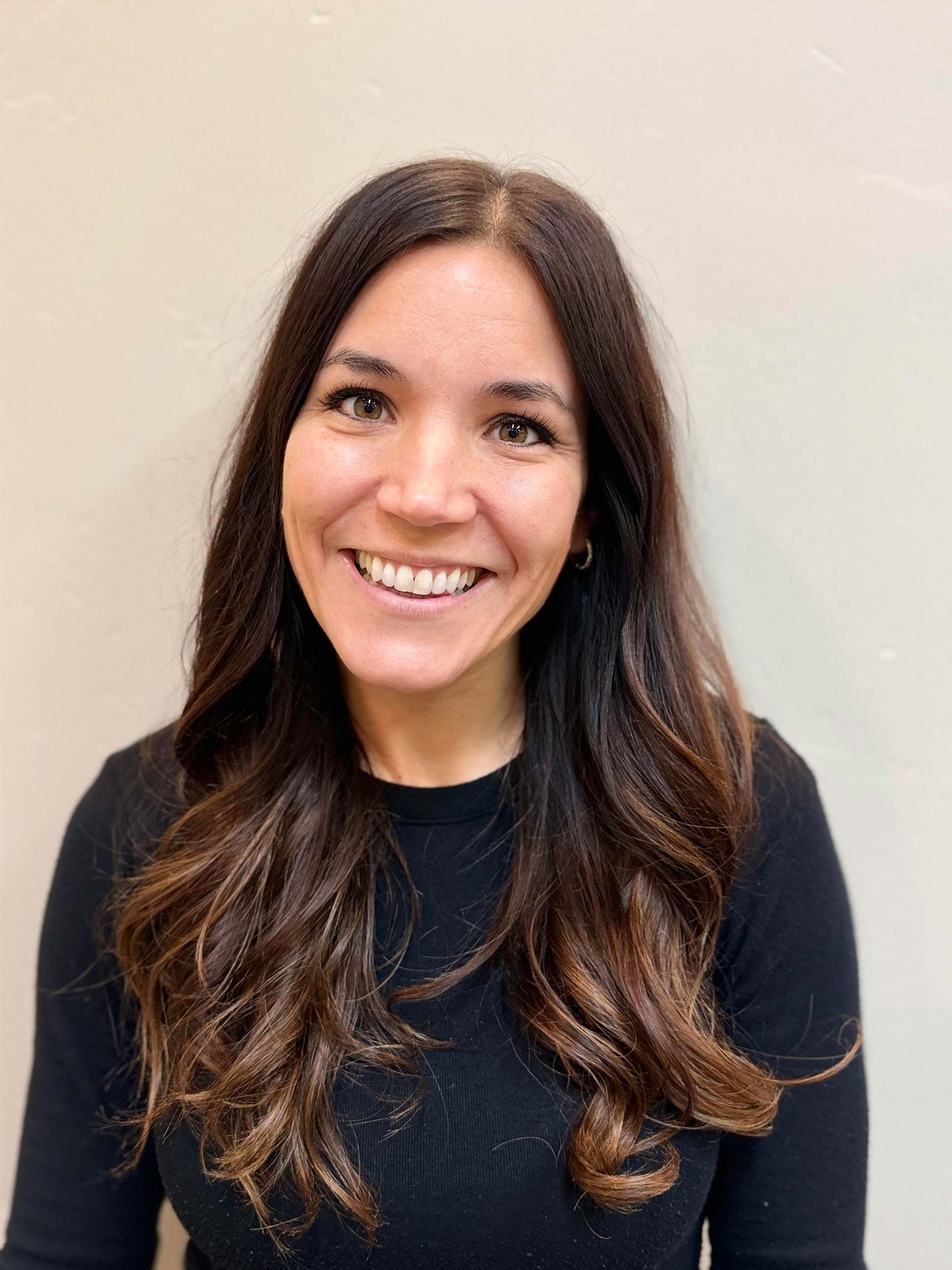 Woman smiling, dark hair, wearing a black top, against a neutral-colored wall.
