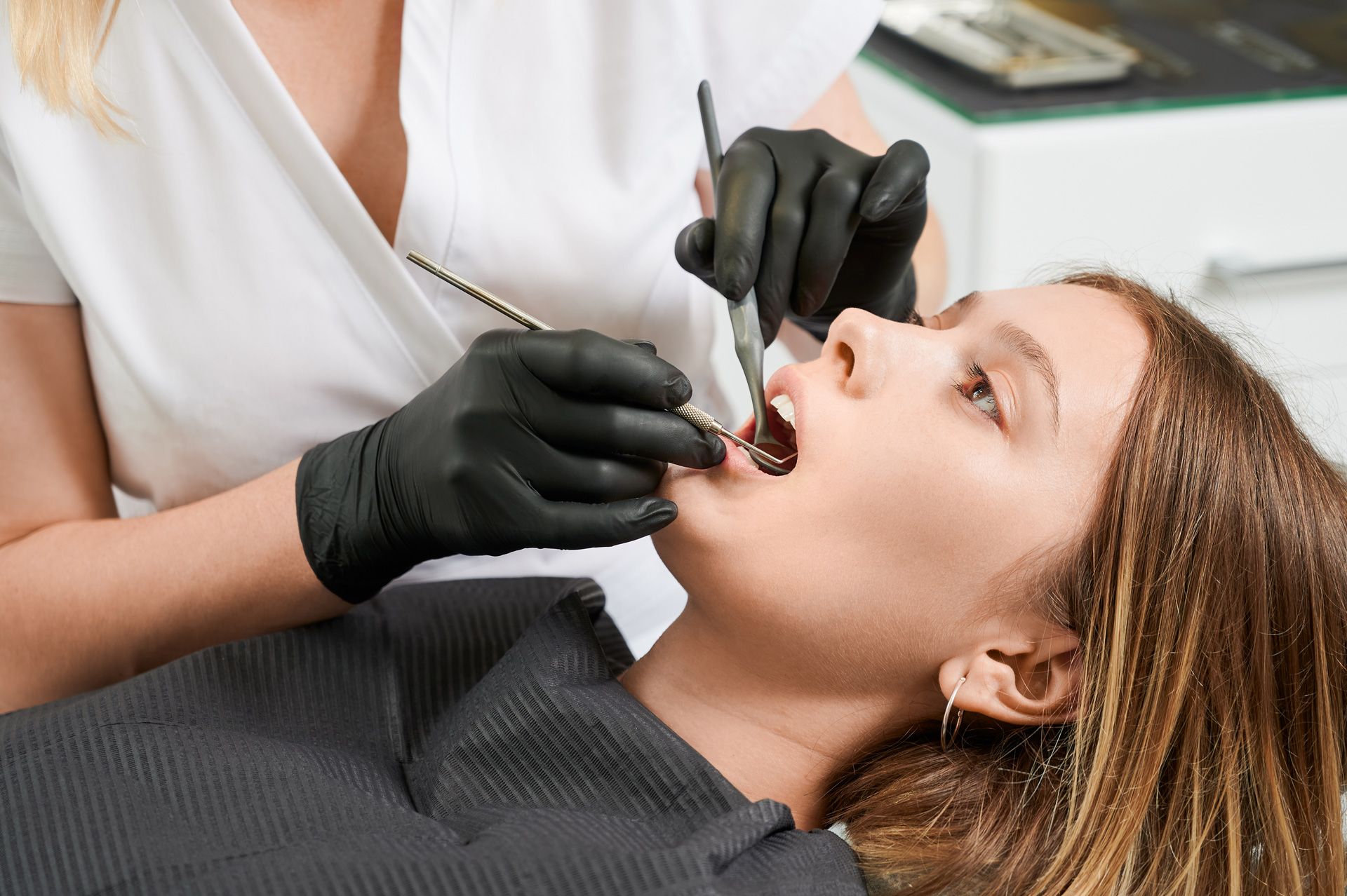 Dentist examining a patient's teeth. The patient's mouth is open. Dentist wears black gloves, white top, setting is a dentist's office.