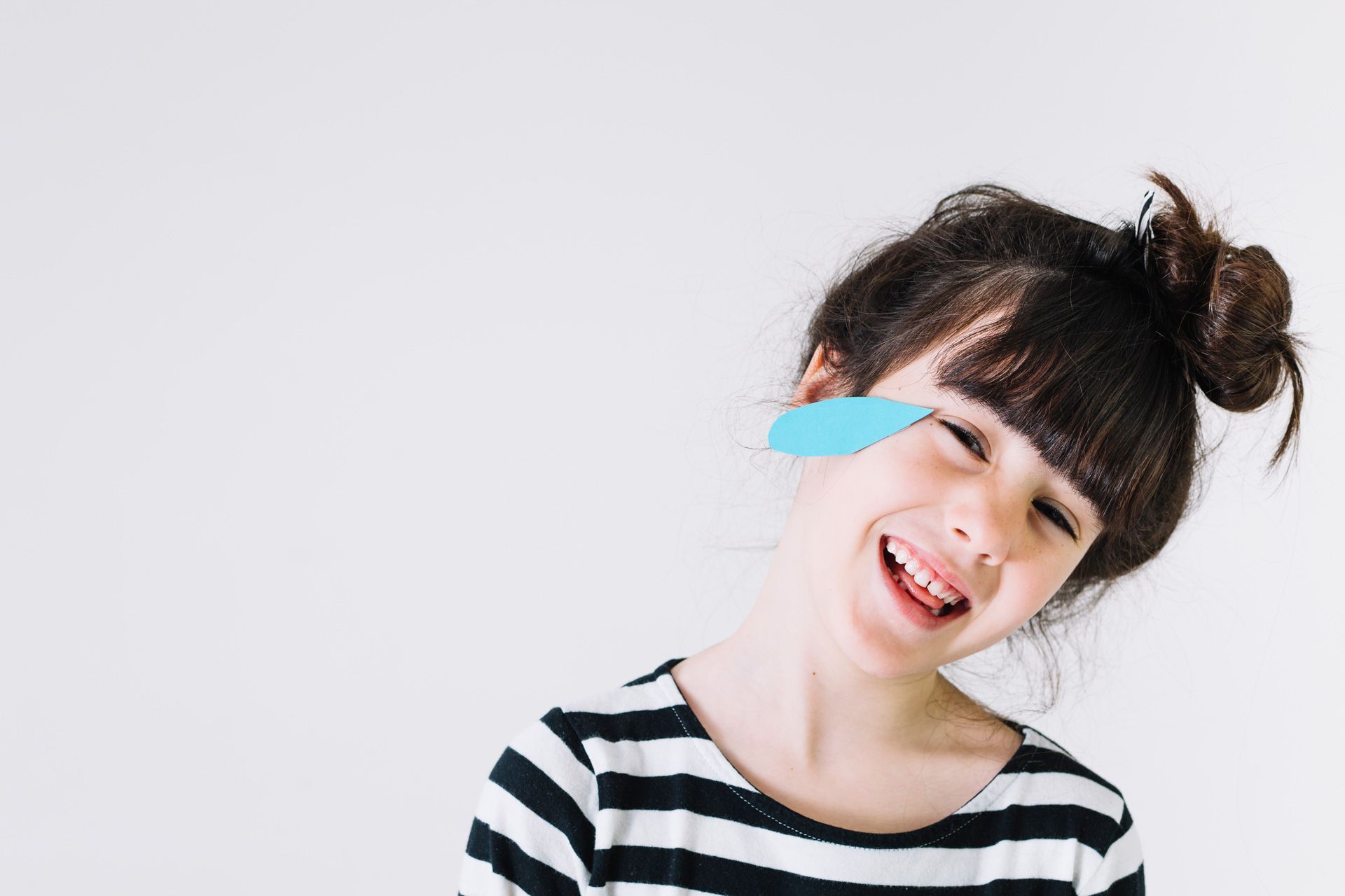 Girl with a paper feather over one eye, smiling broadly, wearing a striped shirt.