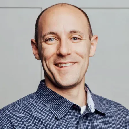 Smiling man with blue eyes in a blue patterned shirt, set against a neutral background.