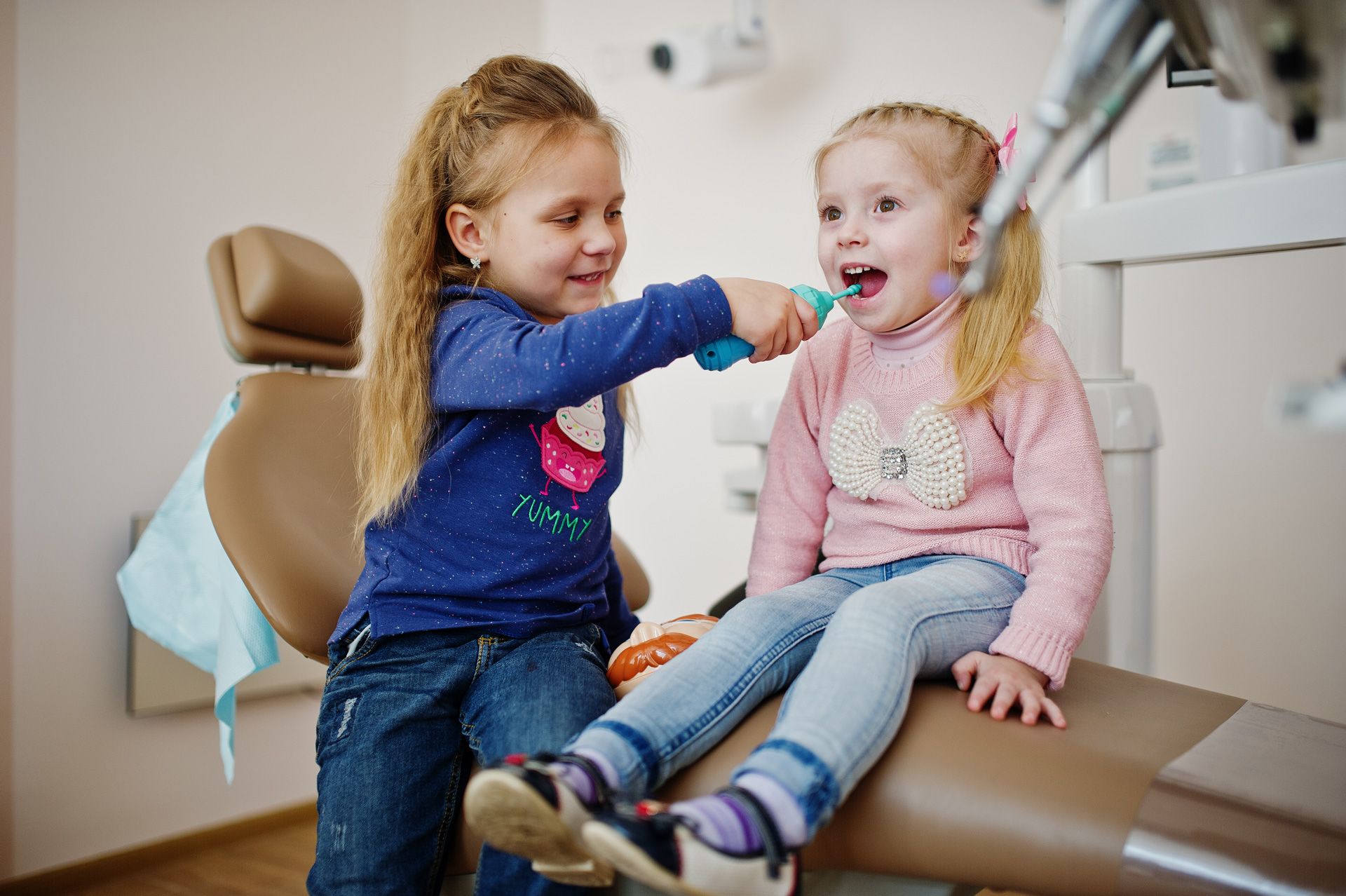 Two girls playing dentist: one pretends to brush the other's teeth in a dentist's chair.