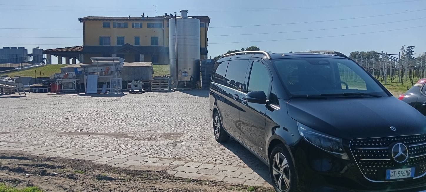 Un furgone Mercedes-Benz nero parcheggiato su un piazzale ghiaioso di fronte a un edificio giallo e a grandi serbatoi metallici industriali.