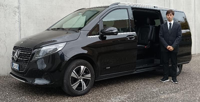 Un autista in giacca e cravatta è in piedi accanto a un minivan Mercedes-Benz nero con la porta scorrevole aperta contro un muro di cemento.