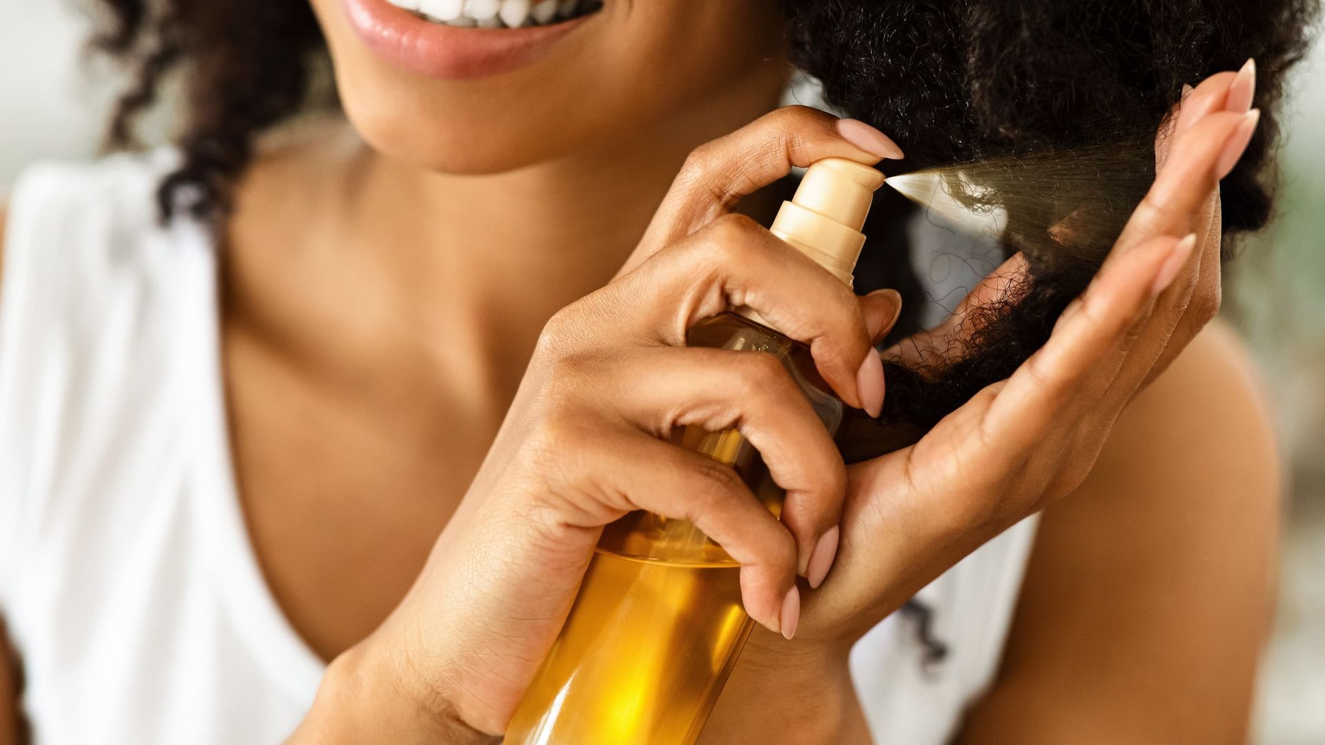 A person sprays a golden hair oil mist onto their dark, curly hair.