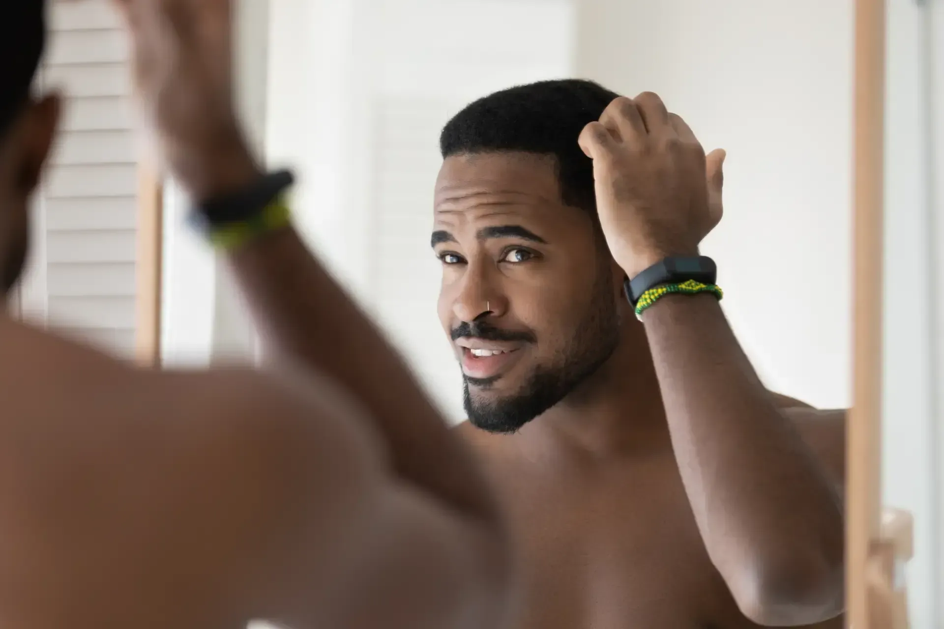 Man adjusting his hair in a bathroom mirror, wearing bracelets and smiling slightly.