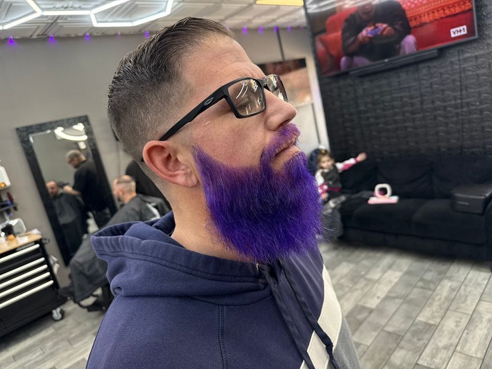 A man with a purple beard is getting his hair cut at a barber shop.