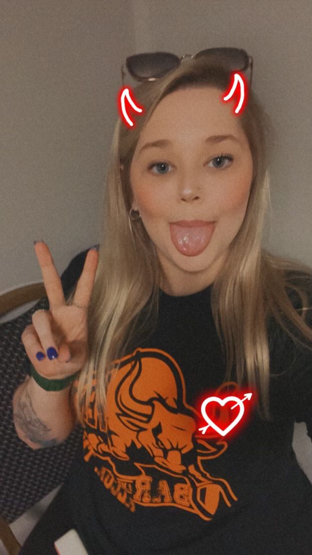 A woman wearing a black shirt with a bull on it is making a peace sign and sticking her tongue out.