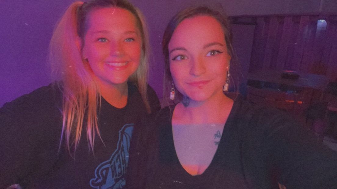 Two women are posing for a picture in front of a purple background.