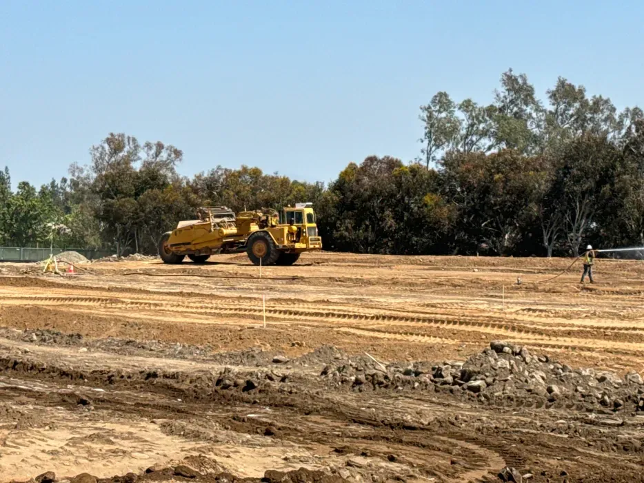 Construction site with earthmover on brown dirt, trees in background, sunny day.