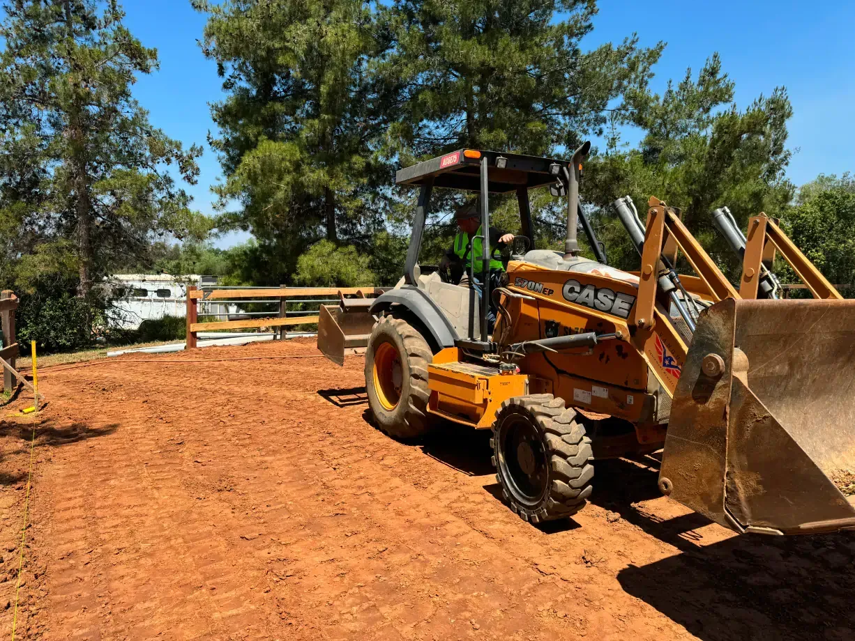 A Case backhoe on a dirt surface is being operated by a person, near a wooden fence.