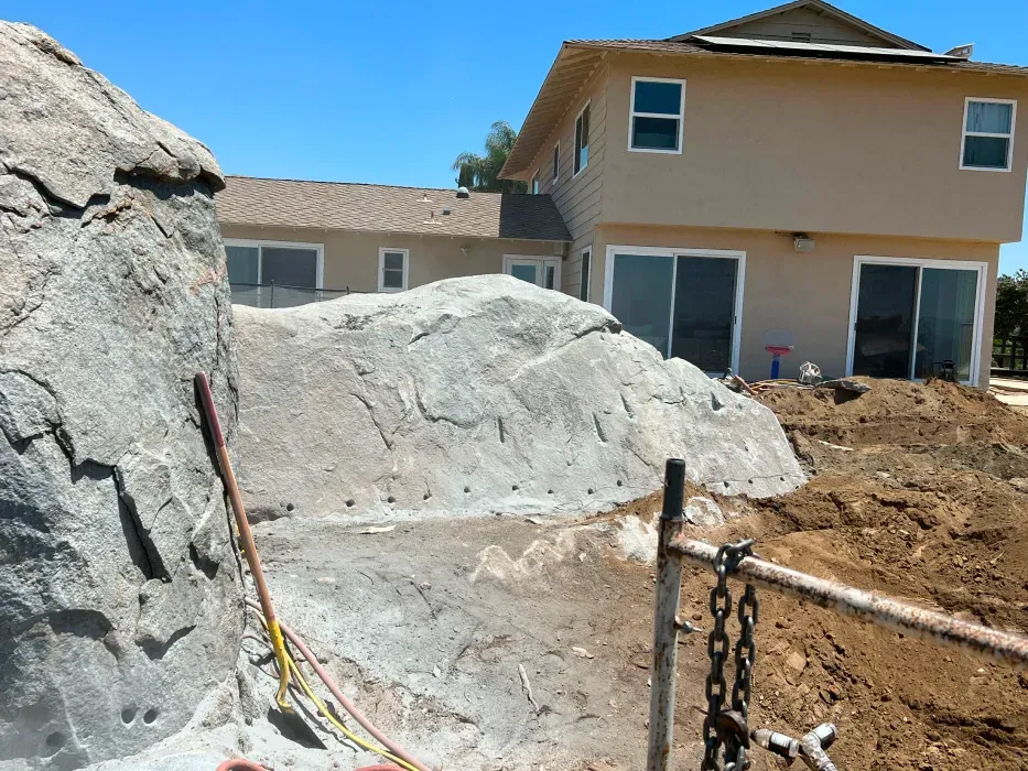 Rock formation next to a two-story beige house under construction; dirt mound and chain fence in the foreground.