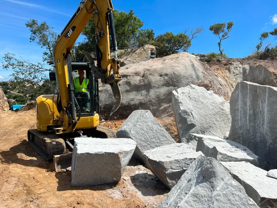 Yellow excavator breaking large gray rocks at a quarry on a sunny day.