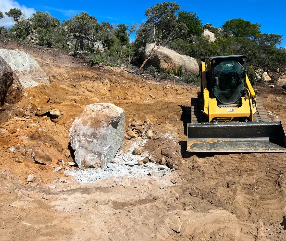 Yellow skid steer excavating around a large rock in a brown, rocky landscape under a blue sky.