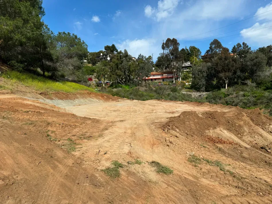 Dirt lot with exposed earth, trees, and a house in the background.