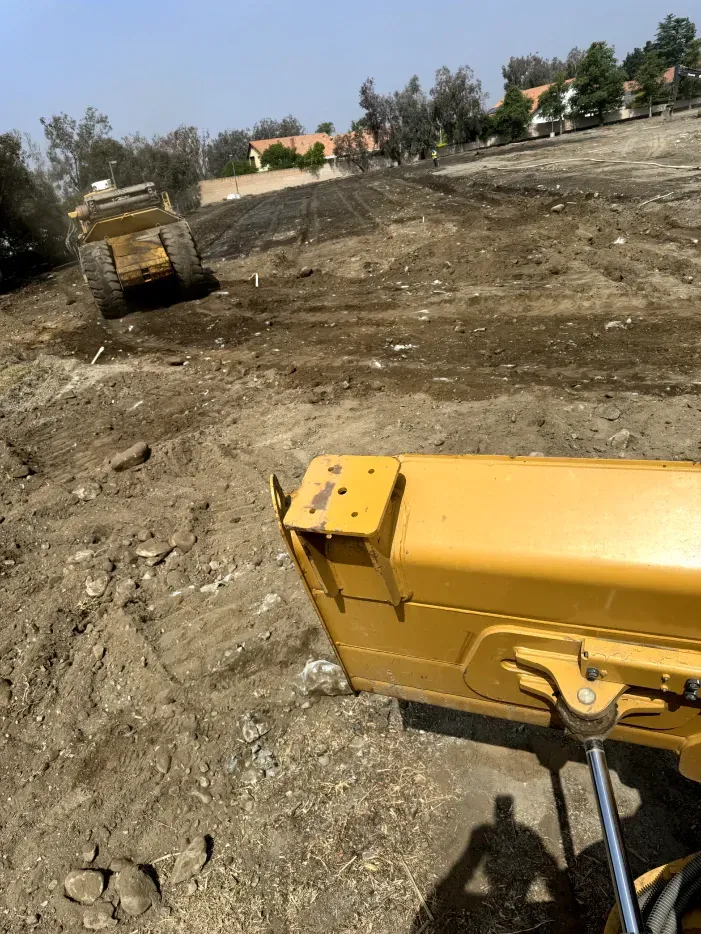 Yellow bulldozer blade in the foreground; another bulldozer levels dirt on a construction site. Sunny day.