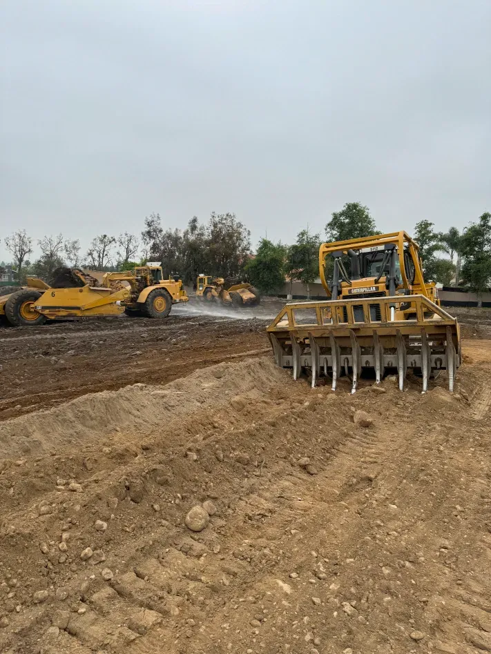 Yellow construction equipment leveling and preparing a brown dirt field under a cloudy sky.