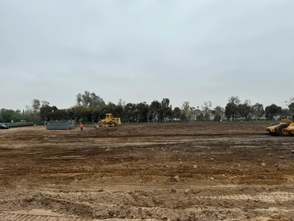 Construction site, dirt field, yellow bulldozers, trees in the background, overcast sky.
