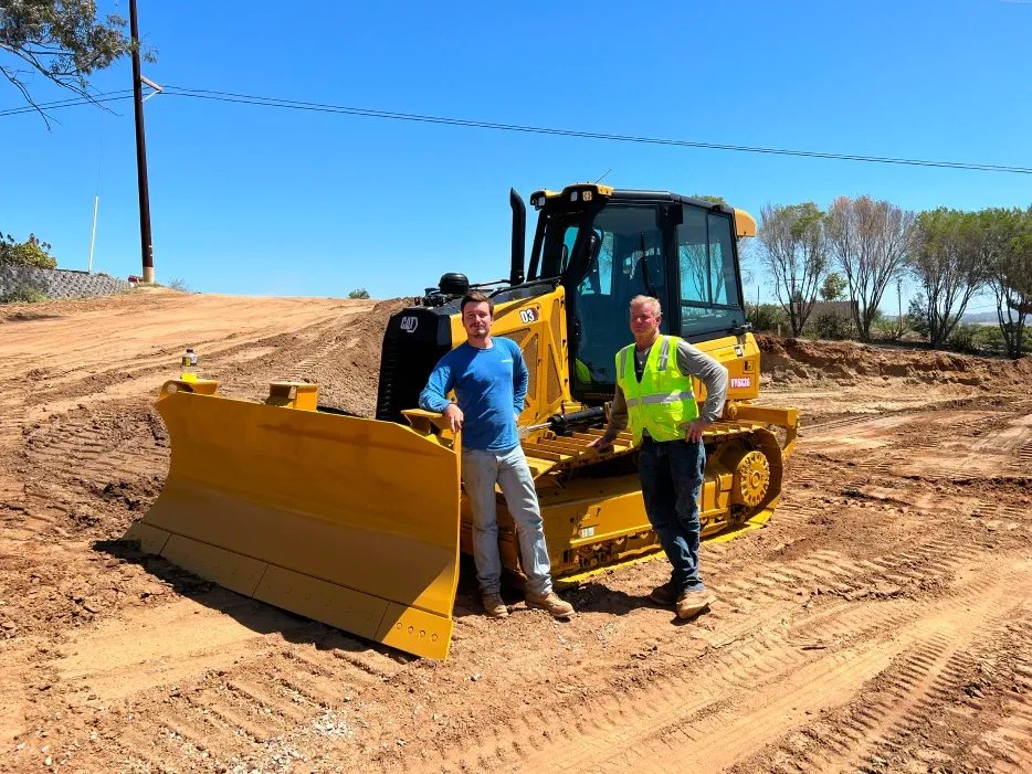 Two men stand near a yellow bulldozer on a construction site under a blue sky.
